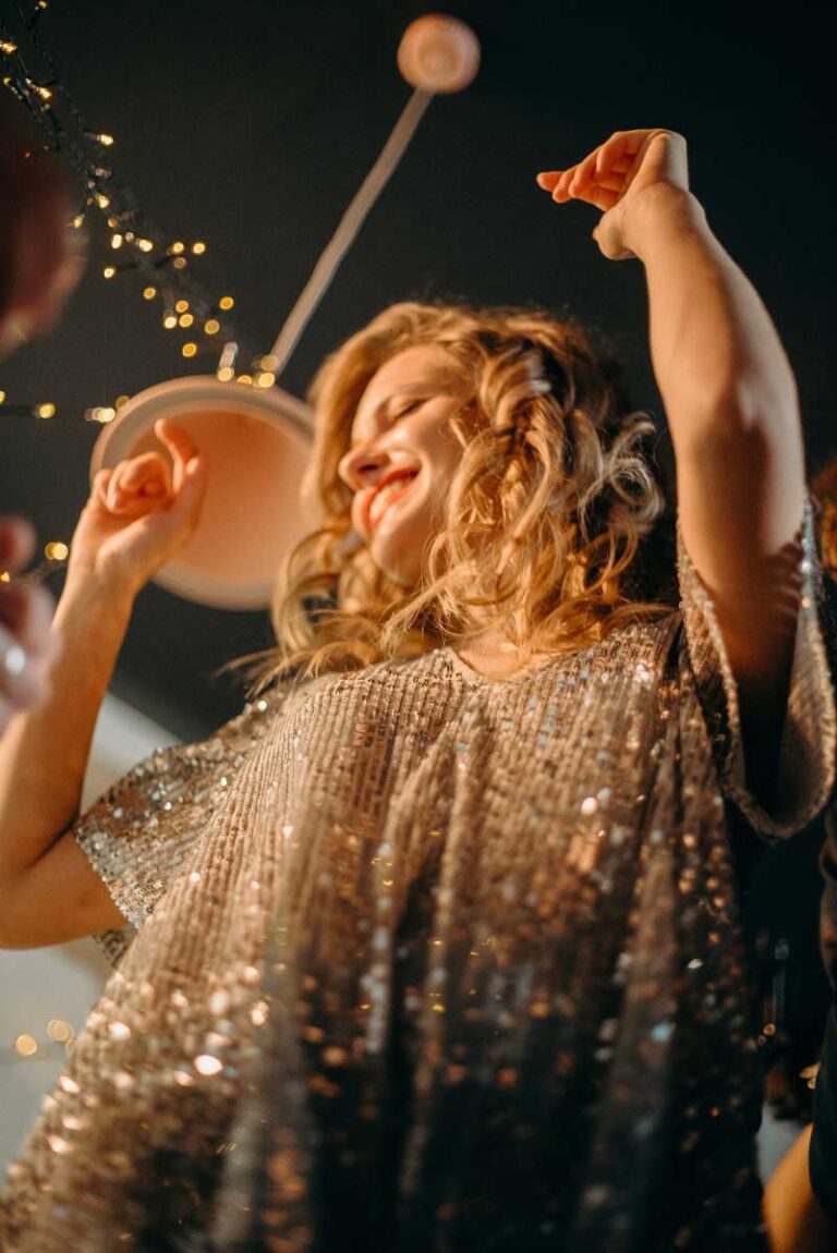 Joyful woman with curly hair dancing at a party, wearing a sparkling dress with lights in the background.