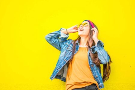 Smiling young woman enjoying music with headphones against yellow wall, wearing denim jacket and colorful headband.