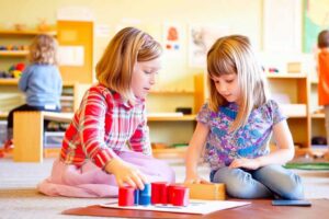 Two young girls sitting on the floor of a bright, colorful Montessori classroom, engaging in a hands-on learning activity.