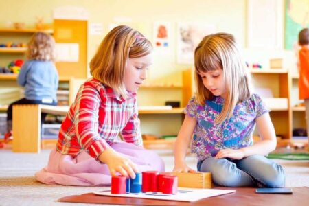 Two young girls sitting on the floor of a bright, colorful Montessori classroom, engaging in a hands-on learning activity.